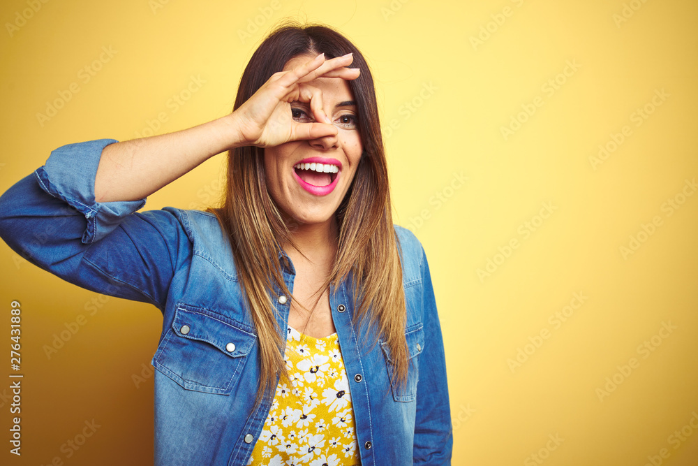 Young beautiful woman standing over yellow isolated background doing ok gesture with hand smiling, eye looking through fingers with happy face. Young beautiful woman standing over yellow isolated background doing ok gesture with hand smiling, eye looking through fingers with happy face.