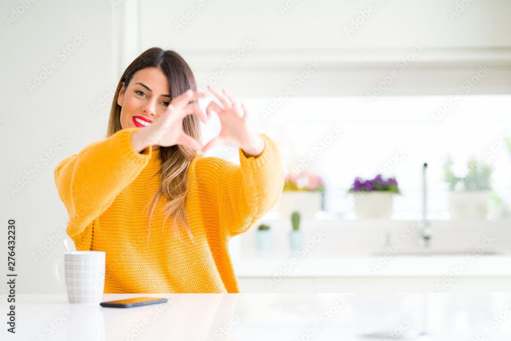 Young beautiful woman drinking a cup of coffee at home smiling in love showing heart symbol and shape with hands. Romantic concept. Young beautiful woman drinking a cup of coffee at home smiling in love showing heart symbol and shape with hands. Romantic concept.