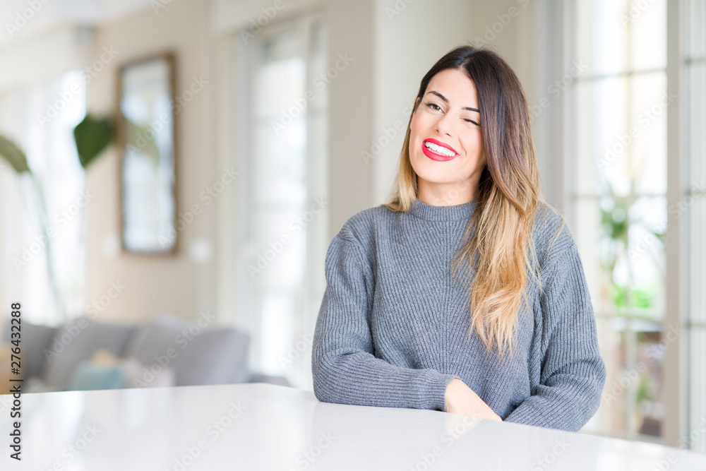 Young beautiful woman wearing winter sweater at home winking looking at the camera with sexy expression, cheerful and happy face. Young beautiful woman wearing winter sweater at home winking looking at the camera with sexy expression, cheerful and happy face.