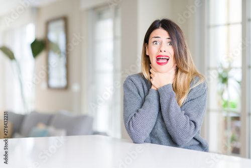 Young beautiful woman wearing winter sweater at home shouting and suffocate because painful strangle. Health problem. Asphyxiate and suicide concept.