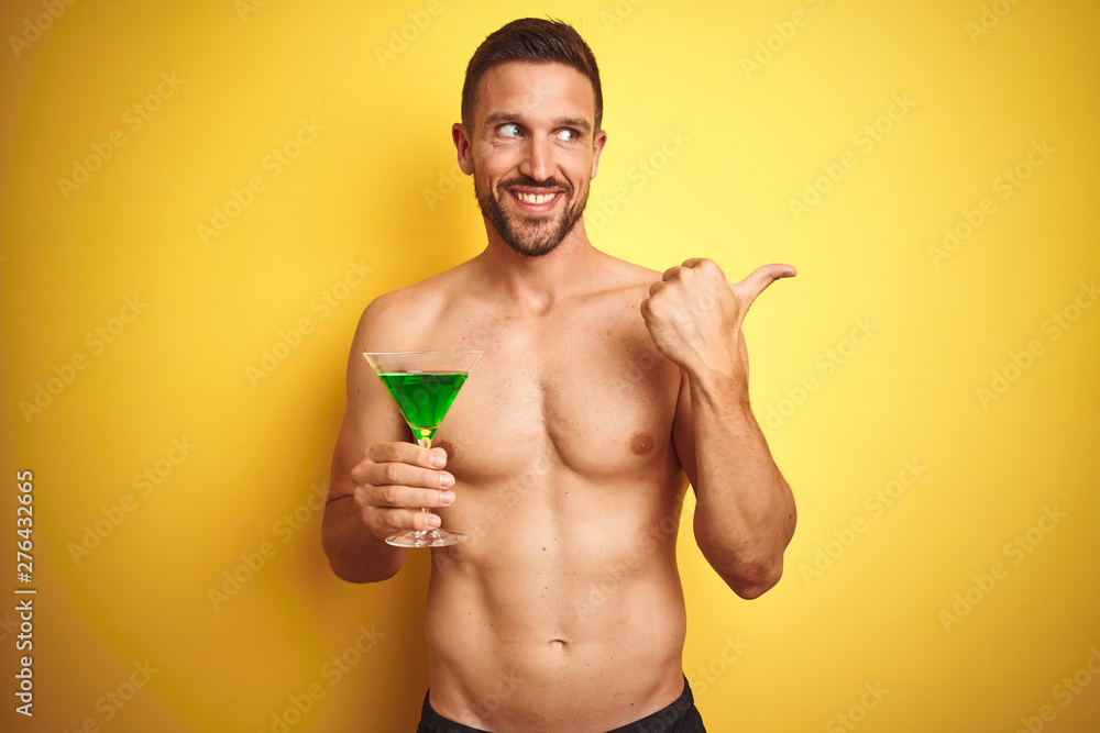 Young handsome shirtless man drinking a summer cocktail over isolated yellow background pointing and showing with thumb up to the side with happy face smiling