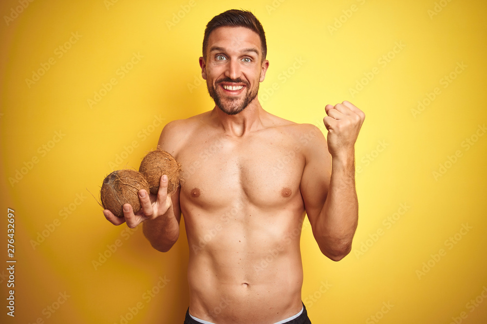Fototapeta premium Young handsome shirtless man holding exotic tropical coconut over isolated yellow background screaming proud and celebrating victory and success very excited, cheering emotion