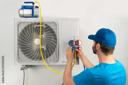 Installation service fix repair maintenance of an air conditioner outdoor unit, by cryogenist technican worker evacuate the system with vacuum pump and manifold gauges in blue shirt baseball cap