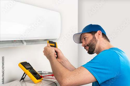 Installation service fix  repair maintenance of an air conditioner indoor unit, by cryogenist technican worker checking the temperature with infrared thermometer in blue shirt baseball cap photo