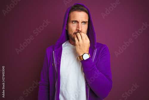 Young fitness man wearing casual sports sweatshirt and hood over purple isolated background looking stressed and nervous with hands on mouth biting nails. Anxiety problem.