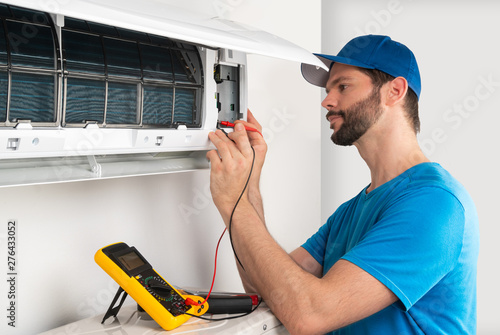Installation service fix repair maintenance of an air conditioner indoor unit, by cryogenist technican worker with multimeter checking electric blue shirt baseball cap photo