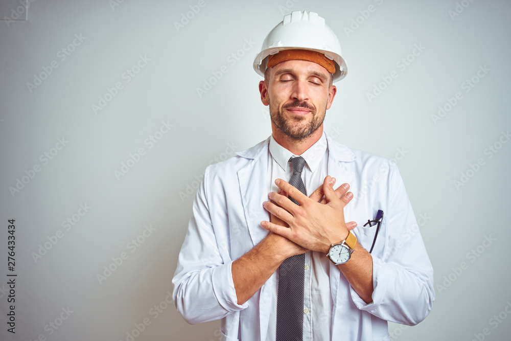 Young handsome engineer man wearing safety helmet over isolated background smiling with hands on chest with closed eyes and grateful gesture on face. Health concept. Young handsome engineer man wearing safety helmet over isolated background smiling with hands on chest with closed eyes and grateful gesture on face. Health concept.