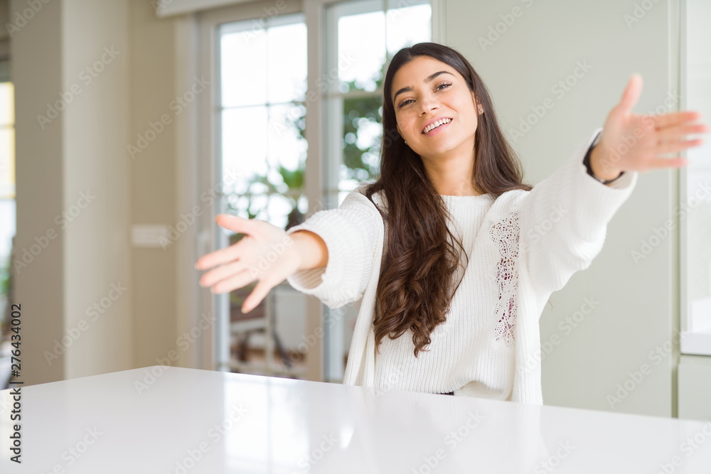 Young beautiful woman at home on white table looking at the camera smiling with open arms for hug. Cheerful expression embracing happiness. Young beautiful woman at home on white table looking at the camera smiling with open arms for hug. Cheerful expression embracing happiness.