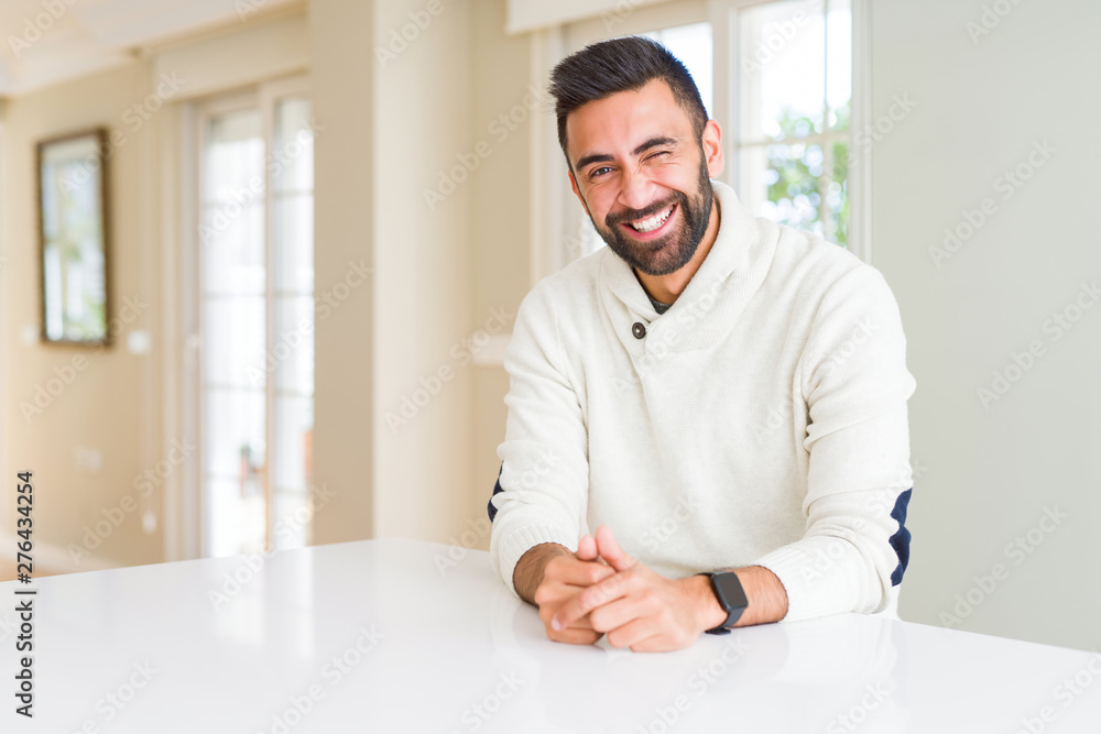 Handsome hispanic man wearing casual white sweater at home winking looking at the camera with sexy expression, cheerful and happy face.