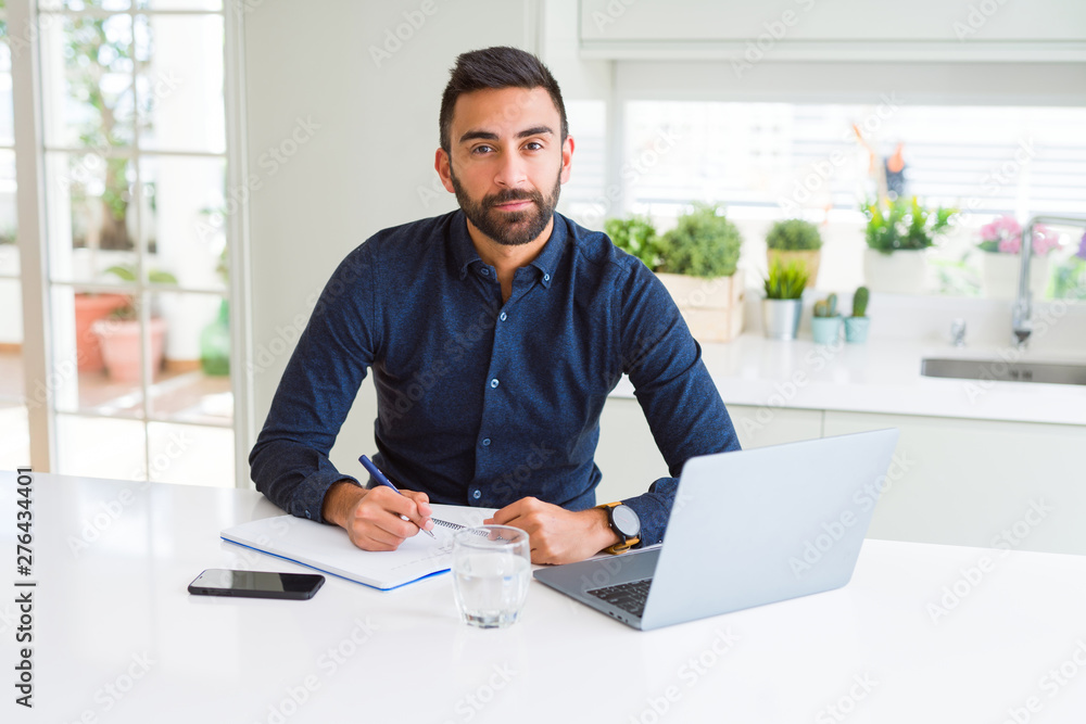 Handsome hispanic man working using computer and writing on a paper with a confident expression on smart face thinking serious Handsome hispanic man working using computer and writing on a paper with a confident expression on smart face thinking serious