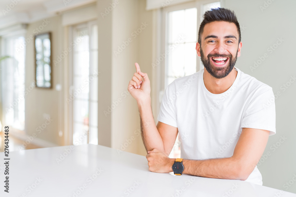 Handsome hispanic man casual white t-shirt at home with a big smile on face, pointing with hand and finger to the side looking at the camera. Handsome hispanic man casual white t-shirt at home with a big smile on face, pointing with hand and finger to the side looking at the camera.