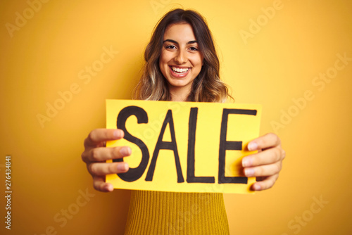 Young beautiful woman holding sale poster over yellow isolated background with a happy face standing and smiling with a confident smile showing teeth