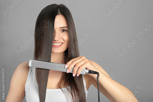 Young woman using hair iron on grey background, space for text