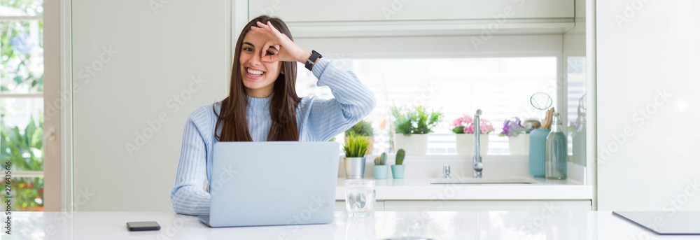 Wide angle picture of beautiful young woman working or studying using laptop with happy face smiling doing ok sign with hand on eye looking through fingers Wide angle picture of beautiful young woman working or studying using laptop with happy face smiling doing ok sign with hand on eye looking through fingers