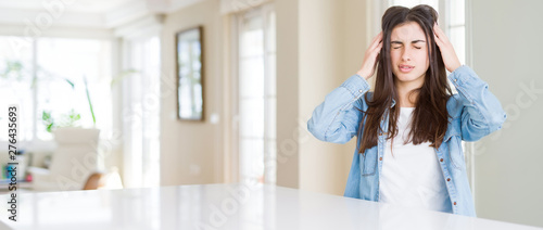 Wide angle picture of beautiful young woman sitting on white table at home with hand on head for pain in head because stress. Suffering migraine.