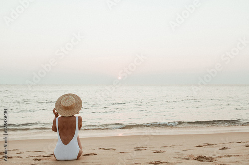 Summer vacation fashion concept. Young, tanned woman wearing a beautiful white swimsuit with a straw hat is sitting and relaxing on tropical beach with white sand and is watching sunset and sea. © Floral Deco