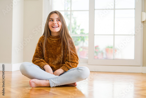 Beautiful young girl kid sitting on the floor at home looking away to side with smile on face, natural expression. Laughing confident.