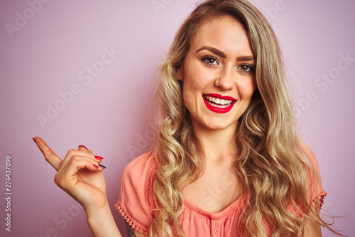 Young beautiful woman wearing t-shirt standing over pink isolated background very happy pointing with hand and finger to the side