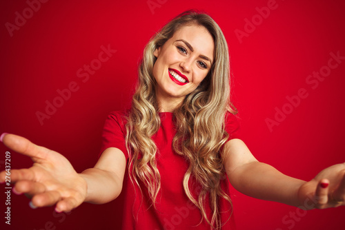 Young beautiful woman wearing basic t-shirt standing over red isolated background looking at the camera smiling with open arms for hug. Cheerful expression embracing happiness.