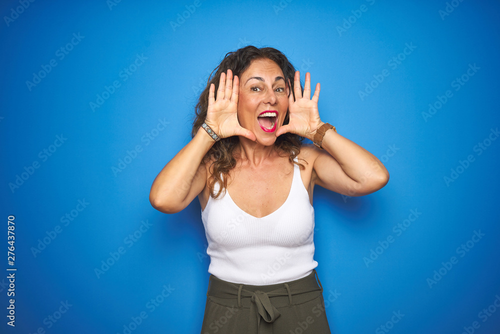 Middle age senior woman with curly hair standing over blue isolated background Smiling cheerful playing peek a boo with hands showing face. Surprised and exited