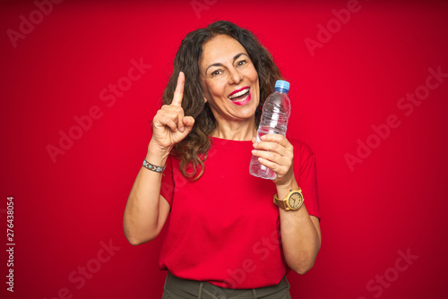 Middle age senior woman holding plastic water bottle over red isolated background surprised with an idea or question pointing finger with happy face, number one
