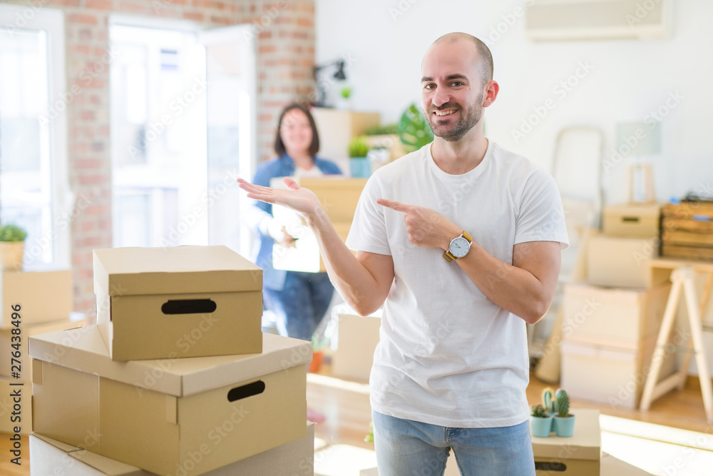 Young couple arround cardboard boxes moving to a new house, bald man standing at home amazed and smiling to the camera while presenting with hand and pointing with finger. Young couple arround cardboard boxes moving to a new house, bald man standing at home amazed and smiling to the camera while presenting with hand and pointing with finger.