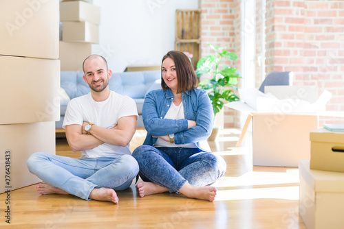 Young couple sitting on the floor arround cardboard boxes moving to a new house happy face smiling with crossed arms looking at the camera. Positive person.