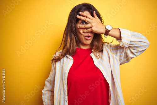 Young beautiful woman wearing red t-shirt and stripes shirt over yellow isolated background peeking in shock covering face and eyes with hand, looking through fingers with embarrassed expression. © Krakenimages.com