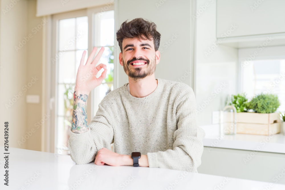 Young man wearing casual sweater sitting on white table smiling positive doing ok sign with hand and fingers. Successful expression.