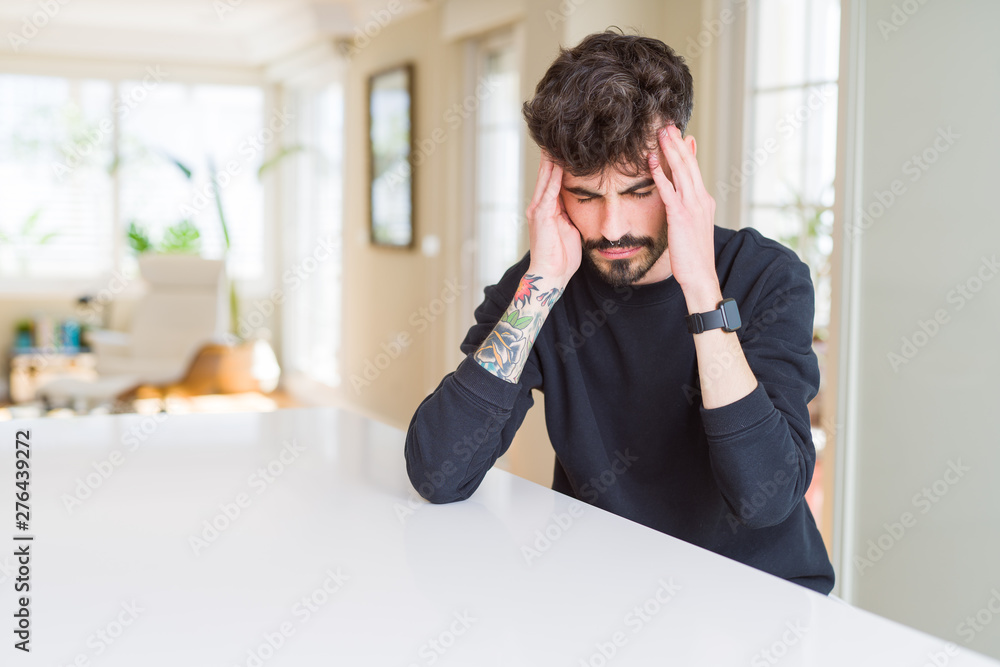 Young man wearing casual sweatshirt sitting on white table suffering from headache desperate and stressed because pain and migraine. Hands on head. Young man wearing casual sweatshirt sitting on white table suffering from headache desperate and stressed because pain and migraine. Hands on head.