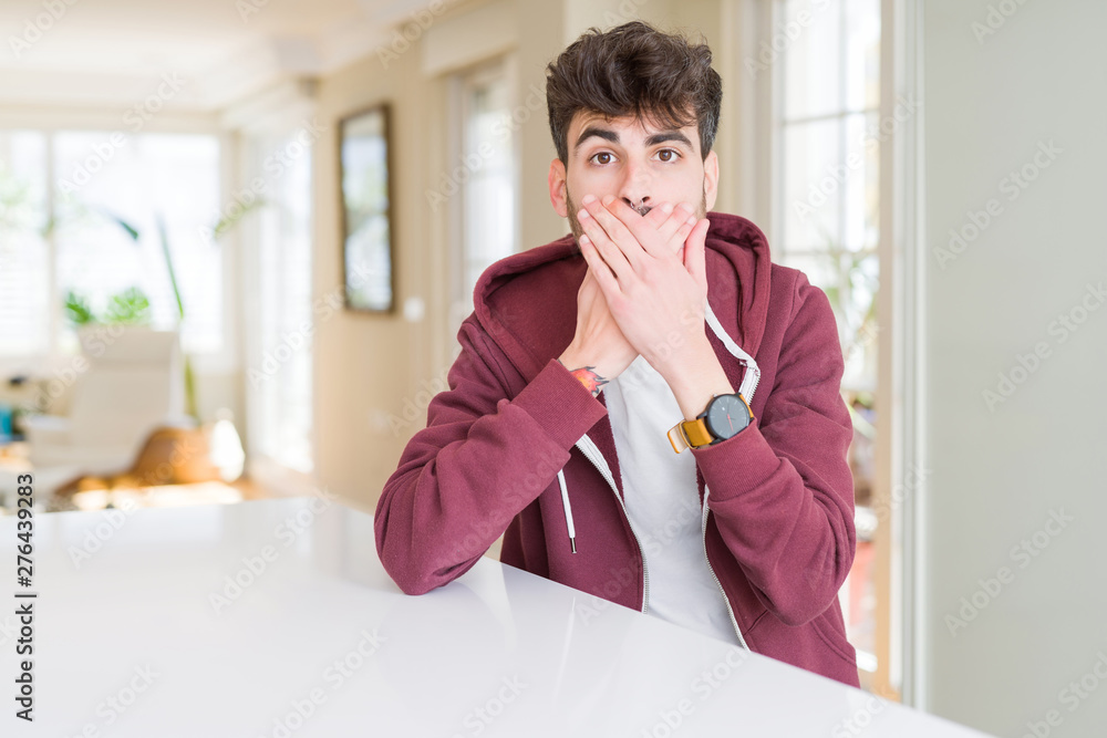 Young man wearing casual sweatshirt sitting on white table shocked covering mouth with hands for mistake. Secret concept. Young man wearing casual sweatshirt sitting on white table shocked covering mouth with hands for mistake. Secret concept.