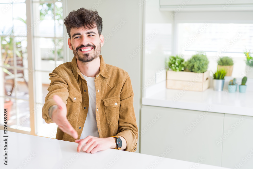 Young man wearing casual jacket sitting on white table smiling friendly offering handshake as greeting and welcoming. Successful business.