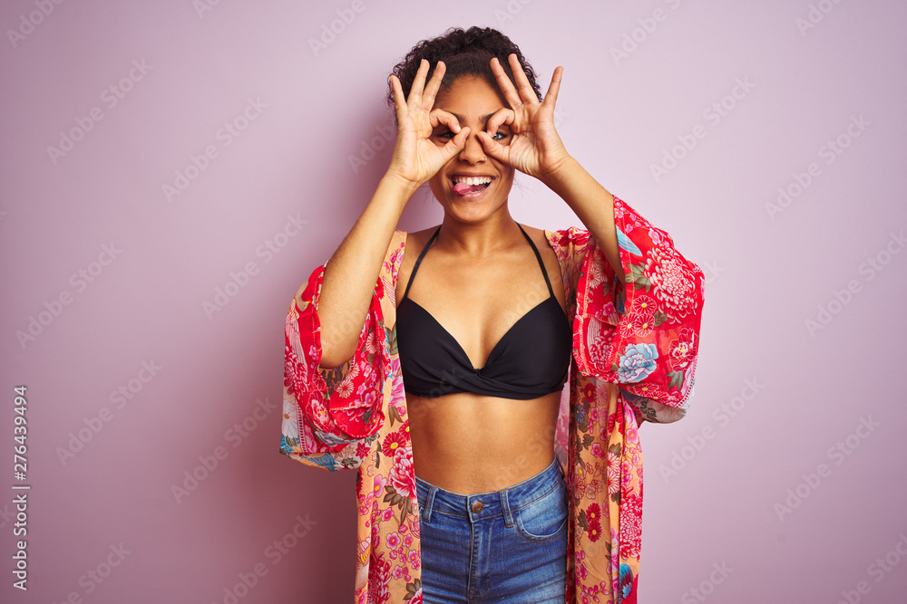 American woman on holiday wearing bikini and colorful dress over isolated pink background doing ok gesture like binoculars sticking tongue out, eyes looking through fingers. Crazy expression. American woman on holiday wearing bikini and colorful dress over isolated pink background doing ok gesture like binoculars sticking tongue out, eyes looking through fingers. Crazy expression.