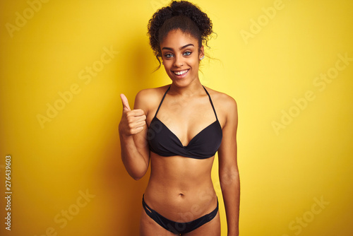 African american woman on vacation wearing bikini standing over isolated yellow background doing happy thumbs up gesture with hand. Approving expression looking at the camera showing success. © Krakenimages.com