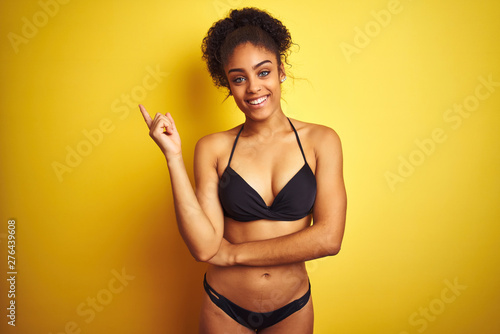 African american woman on vacation wearing bikini standing over isolated yellow background with a big smile on face, pointing with hand finger to the side looking at the camera.