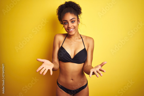 African american woman on vacation wearing bikini standing over isolated yellow background smiling cheerful offering hands giving assistance and acceptance. © Krakenimages.com