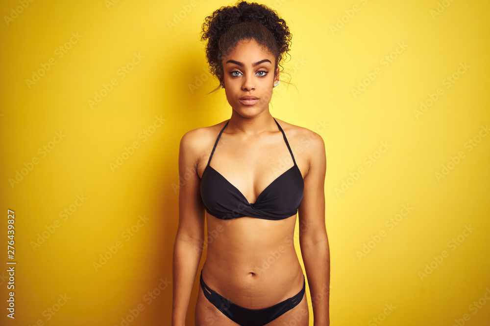 African american woman on vacation wearing bikini standing over isolated yellow background Relaxed with serious expression on face. Simple and natural looking at the camera. African american woman on vacation wearing bikini standing over isolated yellow background Relaxed with serious expression on face. Simple and natural looking at the camera.