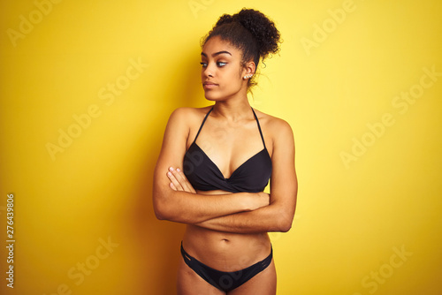 African american woman on vacation wearing bikini standing over isolated yellow background looking to the side with arms crossed convinced and confident