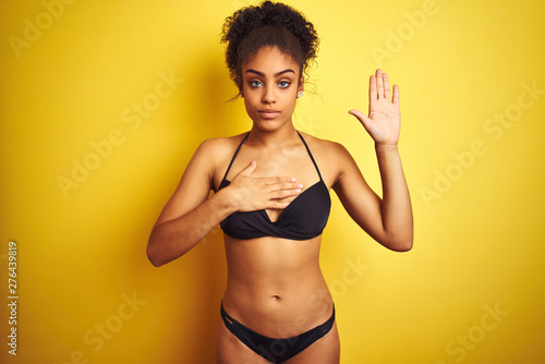 African american woman on vacation wearing bikini standing over isolated yellow background Swearing with hand on chest and open palm, making a loyalty promise oath