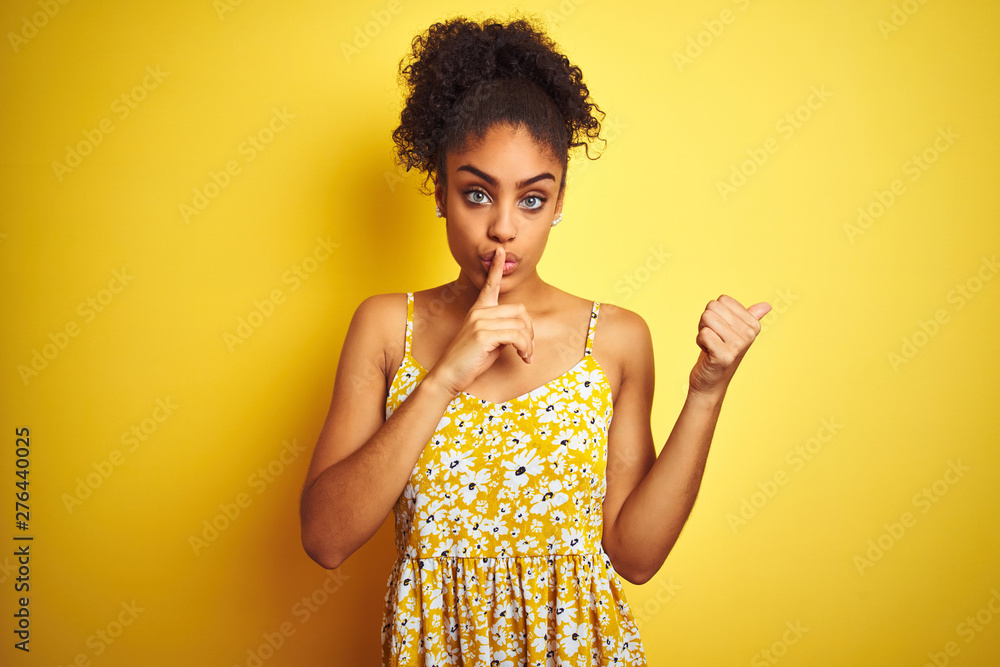 African american woman wearing casual floral dress standing over isolated yellow background asking to be quiet with finger on lips pointing with hand to the side. Silence and secret concept. African american woman wearing casual floral dress standing over isolated yellow background asking to be quiet with finger on lips pointing with hand to the side. Silence and secret concept.