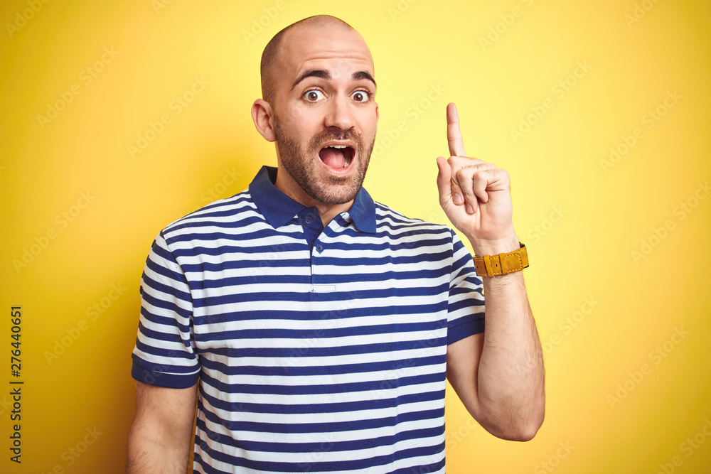 Young bald man with beard wearing casual striped blue t-shirt over yellow isolated background pointing finger up with successful idea. Exited and happy. Number one. Young bald man with beard wearing casual striped blue t-shirt over yellow isolated background pointing finger up with successful idea. Exited and happy. Number one.