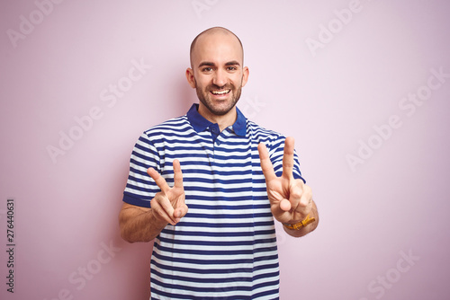Young bald man with beard wearing casual striped blue t-shirt over pink isolated background smiling looking to the camera showing fingers doing victory sign. Number two.