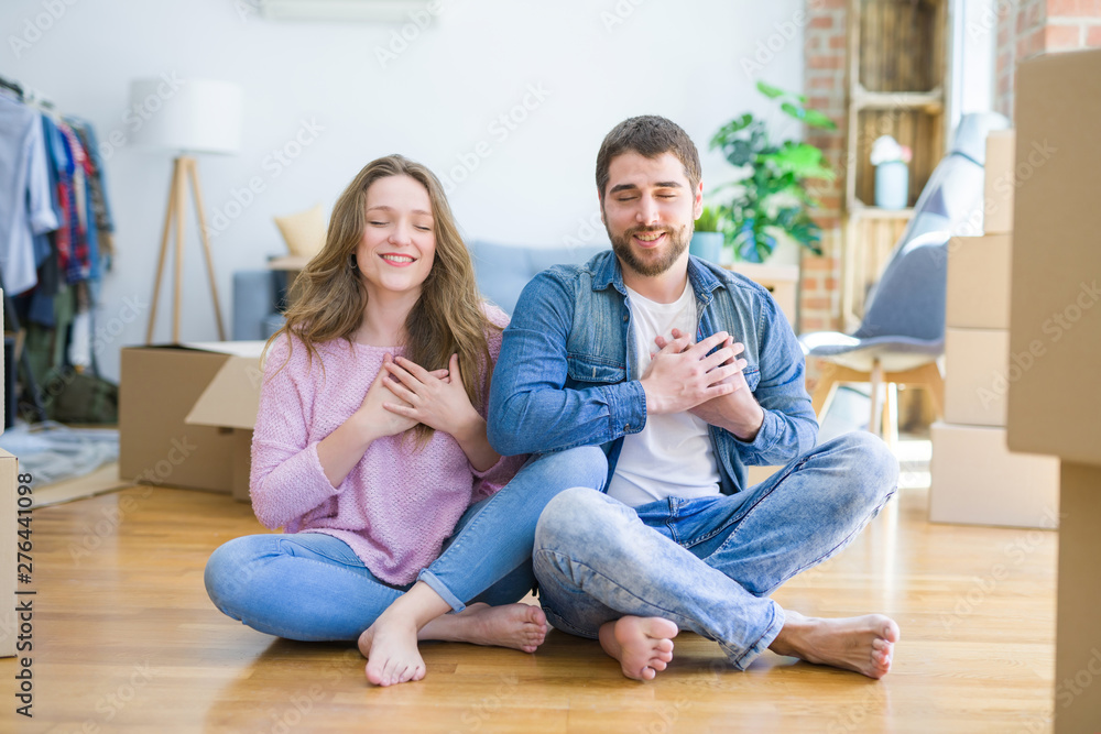 Young beautiful couple moving to a new house sitting on the floor smiling with hands on chest with closed eyes and grateful gesture on face. Health concept. Young beautiful couple moving to a new house sitting on the floor smiling with hands on chest with closed eyes and grateful gesture on face. Health concept.