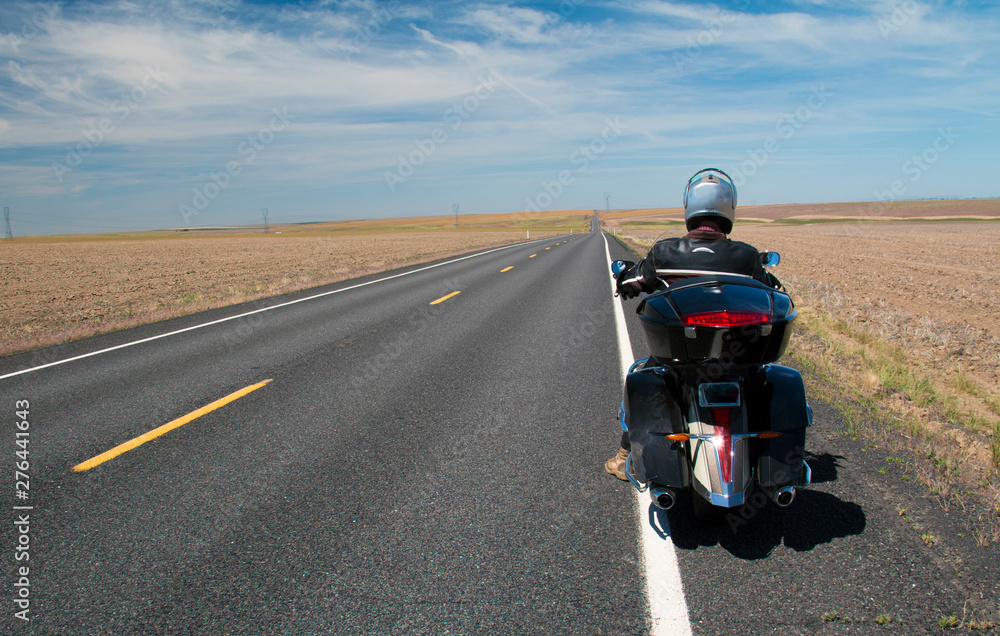 Man resting on motorcycle on roadside with long highway and sky in distance with wispy clouds