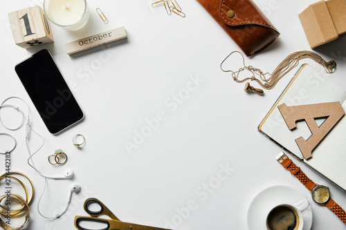 top view of smartphone, notepad, candle, case, earphones, plants, office supplies and coffee with jewelry on white surface