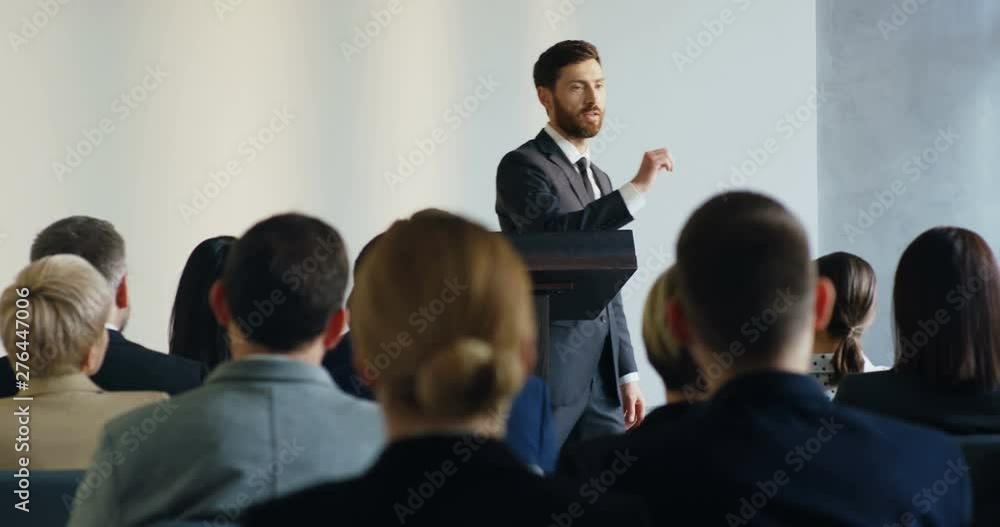 Young Caucasian business trainer walking at the podium and teaching his audience during the conference speech. Young Caucasian business trainer walking at the podium and teaching his audience during the conference speech.