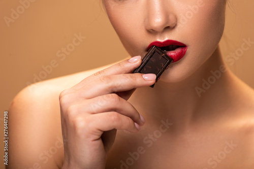 cropped view of naked woman holding chocolate piece near lips isolated on beige