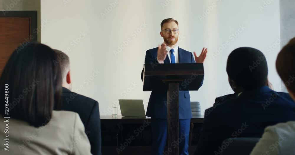 View from among the people in audience on the Caucasian god looking male business coach standing at the lecturn and speaking to the audience. View from among the people in audience on the Caucasian god looking male business coach standing at the lecturn and speaking to the audience.