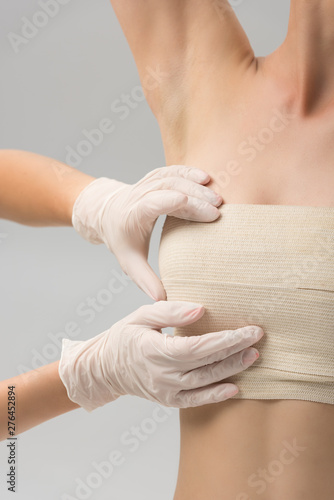 partial view of plastic surgeon in latex gloves and patient in breast bandage isolated on grey