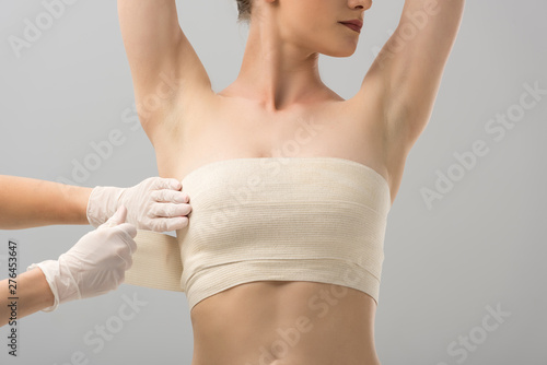 partial view of plastic surgeon in latex gloves and patient in breast bandage isolated on grey photo